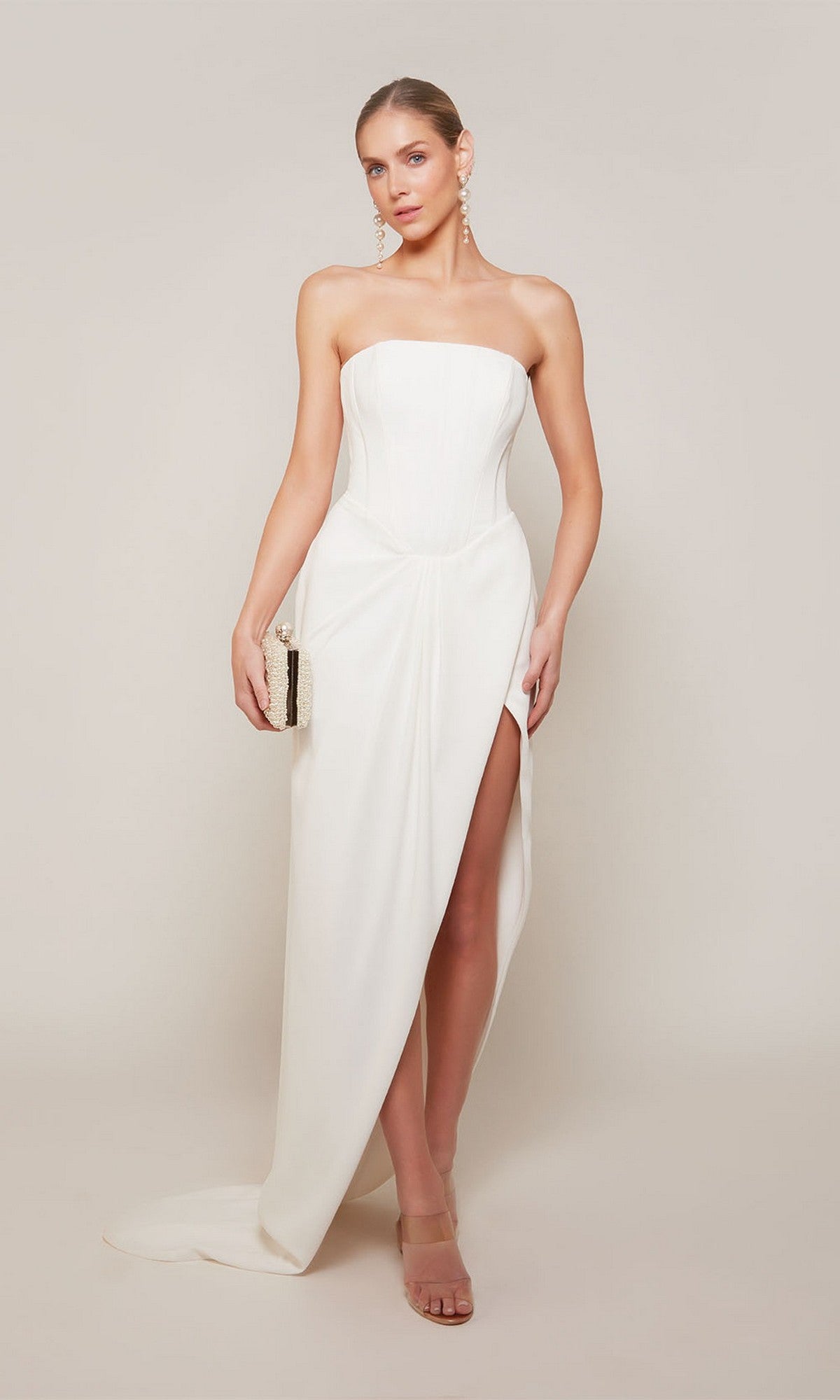 A woman wearing the Alyce 70042 long formal dress with a strapless corset bodice and thigh-high slit sheath skirt, holding a small clutch against a plain light background.
